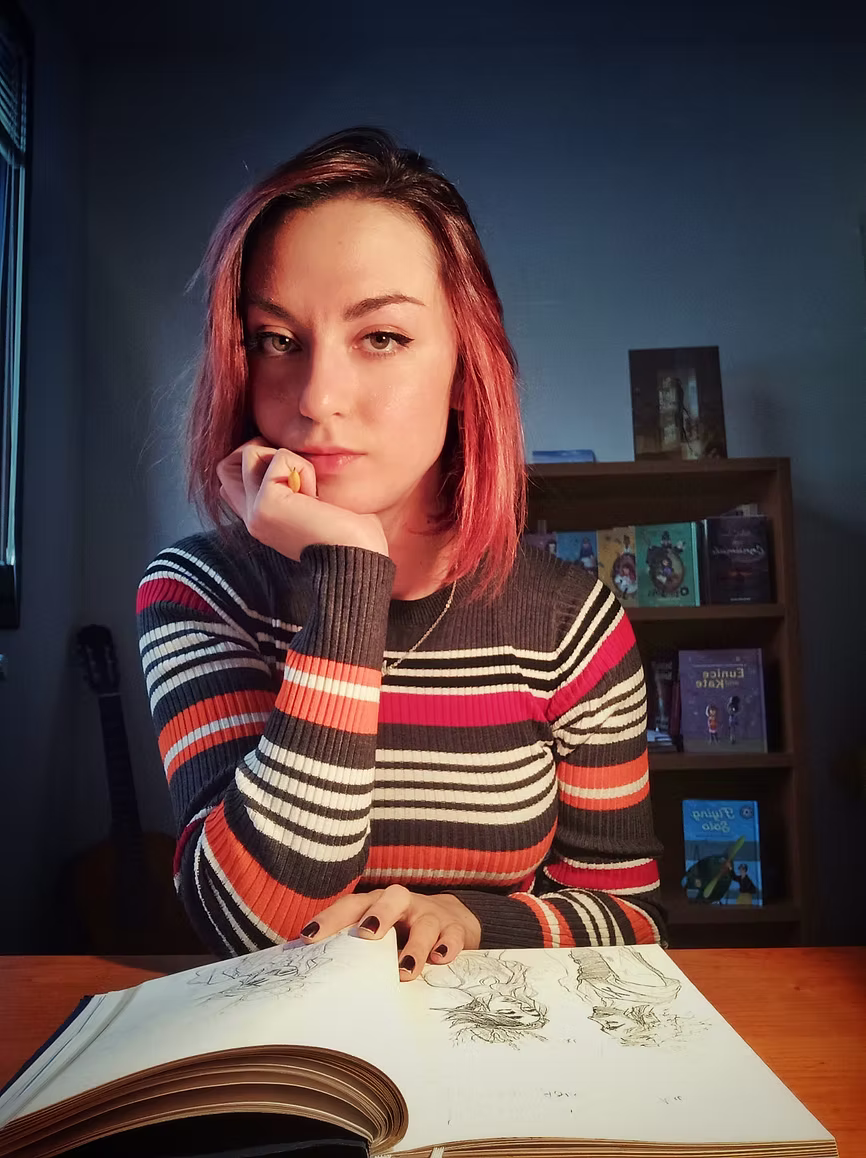 Elena with pink hair wearing a striped sweater, sitting at a table with an open book.